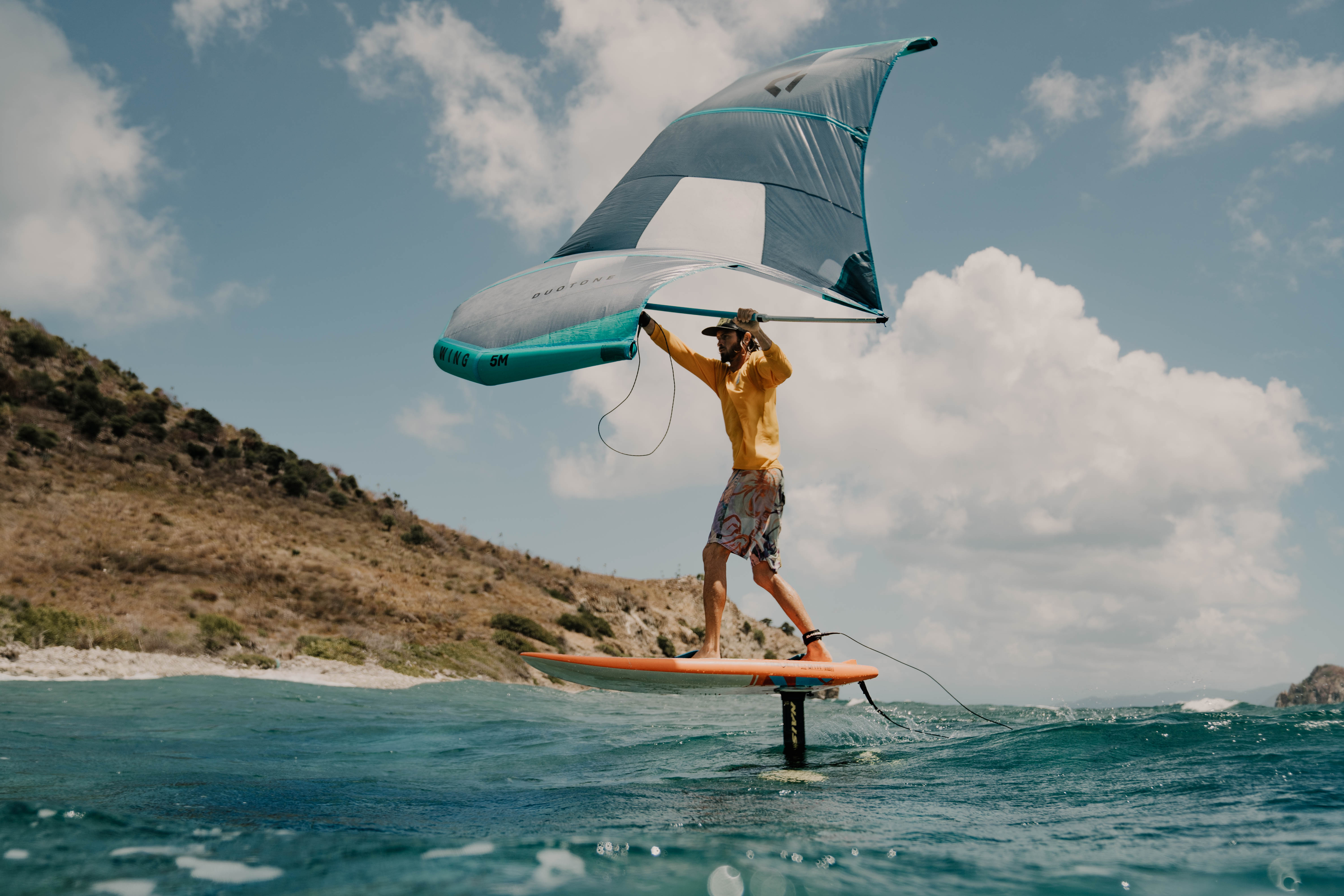 Wing foiler in a yellow top lifting out of turquoise water past a BVI island